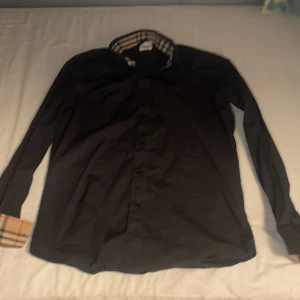 Svart skjorta från Burberry - Säljer en stilren svart skjorta från Burberry i storlek M. Skjortan har långa ärmar och klassiska Burberry-rutor på insidan av kragen och ärmsluten. Den är broderad med Burberrys ikoniska logga på bröstet. Perfekt för både vardag och fest! Inga slitningar på skjortan!