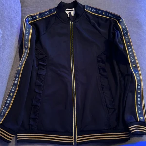 Svart jacka från Scotch & Soda - Säljer en snygg mörkblå jacka från Scotch & Soda. Den har en cool design med gula detaljer och texten 'Scotch Rbelle' längs ärmarna. Jackan har en dragkedja framtill och ribbade muddar vid ärmslut och nederkant. Volanger på framsidan. Perfekt för en avslappnad stil! 