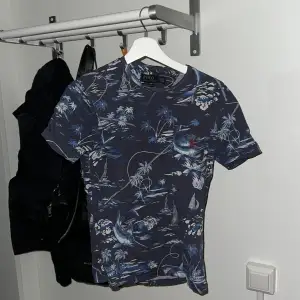 Säljer en snygg blå t-shirt från Polo Ralph Lauren med ett coolt mönster av palmer och båtar. T-shirten är kortärmad och har en slim passform, perfekt för sommarens äventyr. Materialet känns mjukt och skönt mot huden. Passar både till vardags och fest!