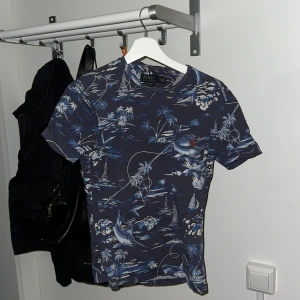 Blå mönstrad t-shirt från Polo Ralph Lauren - Säljer en snygg blå t-shirt från Polo Ralph Lauren med ett coolt mönster av palmer och båtar. T-shirten är kortärmad och har en slim passform, perfekt för sommarens äventyr. Materialet känns mjukt och skönt mot huden. Passar både till vardags och fest!