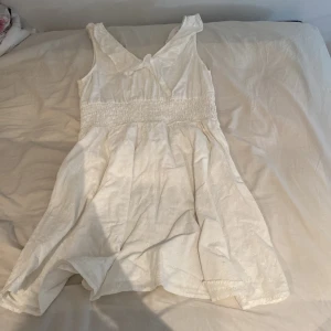 Vit sommarklänning från H&M - Säljer en söt vit sommarklänning från H&M. Klänningen är ärmlös och har en smockdetalj i midjan som ger en fin passform. Den är tillverkad i ett lätt och luftigt material, perfekt för varma sommardagar. Klänningen har en v-ringad hals och en liten volangdetalj vid axlarna. Perfekt för både vardag och fest!