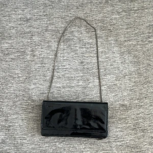 Svart glansig kuvertväska - Säljer en snygg svart glansig kuvertväska med en lång kedjerem. Väskan har en praktisk dragkedja på baksidan och flera fack inuti för kort och andra småsaker. Perfekt för en utekväll eller fest!