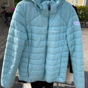 Ljusblå pufferjacka från Everest - Säljer en superfin ljusblå pufferjacka från Everest. Den är perfekt för kyliga dagar med sin quiltade design och mysiga huva. Jackan har en dragkedja framtill och är i ett snyggt och praktiskt material. Perfekt för både höst och vår!