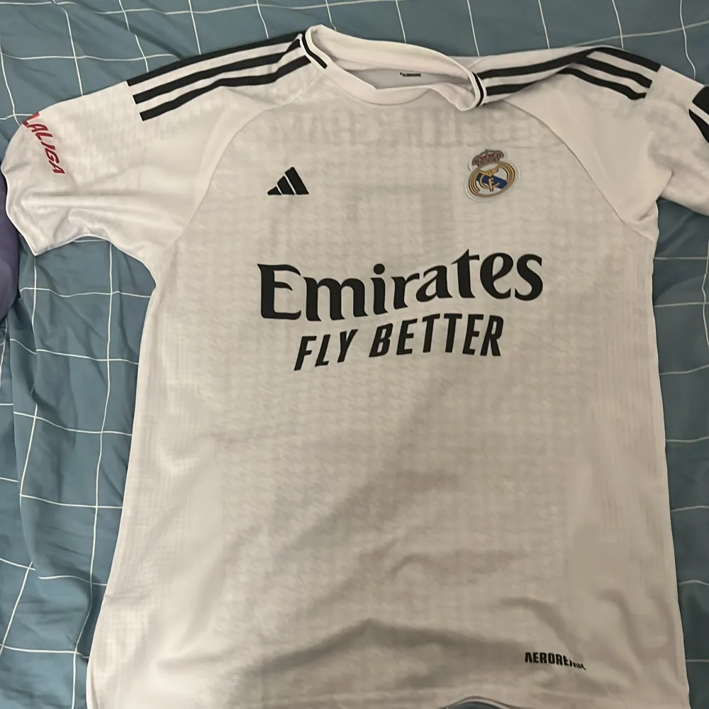 Säljer en vit Real Madrid fotbollströja från Adidas i storlek L. Tröjan har korta ärmar och är i Climacool-material för bra ventilation. Den har klubbens emblem på bröstet och sponsorloggan 'Emirates Fly Better' tryckt på framsidan. På baksidan står det 'Bellingham' och nummer 5. Perfekt för alla Real Madrid-fans!                                 Den är helt sprillans ny. Den va för stor och hade redan dragit av prislappen så kunde inte göra en retur. har inte ens andvänds . T-shirts.