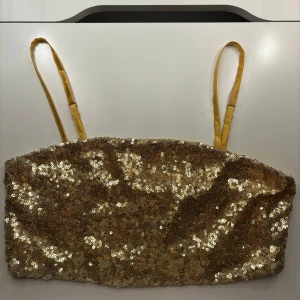 Guldig paljettopp från NA-KD - Säljer en superglittrig guldig paljettopp från NA-KD. Den är axelbandslös med smala axelband och perfekt för festliga tillfällen. Toppen är i storlek 34. Perfekt för nyår eller en utekväll!