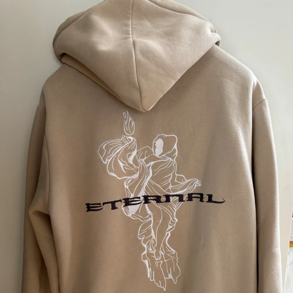 Säljer en snygg beige hoodie med texten 'Eternal' tryckt på både framsidan och baksidan. Den har en cool design med en konstnärlig illustration på ryggen. Hoodien är från HM. Tröjor & Koftor.