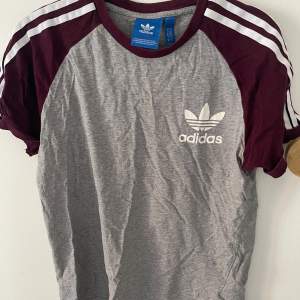 Säljer en snygg grå och vinröd t-shirt från Adidas. Den har korta ärmar och ett klassiskt Adidas-tryck på bröstet. Perfekt för träning eller vardagsbruk. T-shirten är i bomull och har en normal passform. krympt i tvätten och kanske lämpar sig bättre för xs. Köp direkt eller ge prisförslag 💛