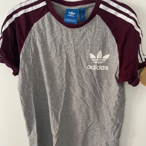 Grå och vinröd t-shirt från Adidas - Säljer en snygg grå och vinröd t-shirt från Adidas. Den har korta ärmar och ett klassiskt Adidas-tryck på bröstet. Perfekt för träning eller vardagsbruk. T-shirten är i bomull och har en normal passform. krympt i tvätten och kanske lämpar sig bättre för xs. Köp direkt eller ge prisförslag 💛