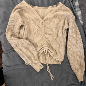 Beige stickad tröja med knytning - Säljer en superfin beige stickad tröja med v-ringning och knytning framtill. Den har långa ärmar och är perfekt för höst och vinter. Tröjan är både mysig och stilren, perfekt för att hålla sig varm och snygg!