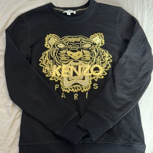 Svart tröja från Kenzo med guldtryck - Säljer en snygg svart tröja från Kenzo med ett coolt guldtryck av en tiger på framsidan. Tröjan har långa ärmar och är perfekt för både vardag och fest. Den är i storlek S och i bra skick. Passar perfekt till jeans eller kjol för en trendig look!
