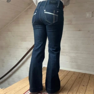 Mörkblå bootcut jeans - Säljer ett par snygga mörkblå bootcut jeans med hög midja. De har coola detaljer med knappar och en klassisk femficksdesign. Perfekta för både vardag och fest!