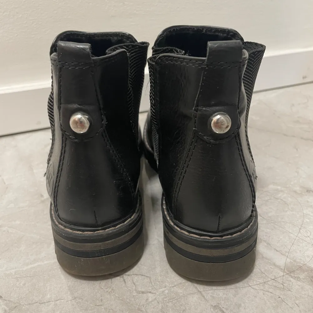 Säljer ett par snygga svarta Chelsea boots från Marco Tozzi. De är i skinn och har en elastisk panel på sidan för enkel på- och avtagning. Skorna har en rund tå och en liten dekorativ knappdetalj på baksidan. Perfekta för höst och vinter!. Skor.