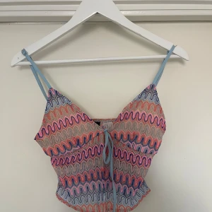 Färgglad topp med vågigt mönster - Säljer en superfin och färgglad topp med vågigt mönster i blått, rosa och beige. Toppen har smala axelband och en söt knytning framtill. Perfekt för sommardagar eller en festlig kväll!