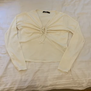 Vit långärmad topp med knytning från Bershka - Säljer en superfin vit långärmad topp från Bershka. Den har en snygg knytning framtill som ger en cool detalj och lite extra stil. Perfekt för både vardag och fest!