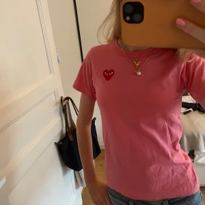 Rosa t-shirt från Comme des Garçons - Säljer en superfin rosa t-shirt från Comme des Garçons med det ikoniska röda hjärtat på bröstet. Den är i storlek XS och är perfekt för en casual look. T-shirten är i bomull och har korta ärmar. Perfekt för sommaren!