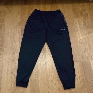 Svarta mjukisbyxor från Puma - Säljer ett par svarta mjukisbyxor från Puma. De har en elastisk midja och muddar vid bensluten för en bekväm passform. På sidorna finns Puma-loggan i grått. Perfekta för en avslappnad dag eller träning.