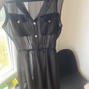 Svart ärmlös klänning - Säljer en svart ärmlös klänning med knappar framtill. Den har en transparent look och två bröstfickor med knappar. Perfekt för en somrig dag eller en festkväll!