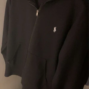 Svart hoodie med dragkedja - Säljer en svart hoodie med dragkedja framtill. Den har en klassisk design med en liten logga på bröstet och en bekväm huva. Perfekt för en avslappnad stil eller för att hålla sig varm under kyliga dagar.