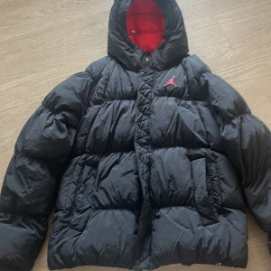Svart pufferjacka från Jordan - Säljer en snygg och varm svart pufferjacka från Jordan. Den har en cool röd logga på bröstet och en dragkedja med Jordan-detaljer. Insidan av huvan är röd, vilket ger en schysst kontrast. Perfekt för kalla vinterdagar!