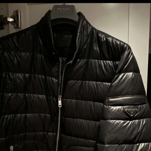 Svart quiltad jacka från Prada - Säljer en snygg svart quiltad jacka från Prada. Den har en stilren design med dragkedja och det ikoniska Prada-emblemet på ärmen. Perfekt för både höst och vinter, och håller dig varm med sin pufferstil. Jackan är i nyskick och kostade 17 000 kr ny 