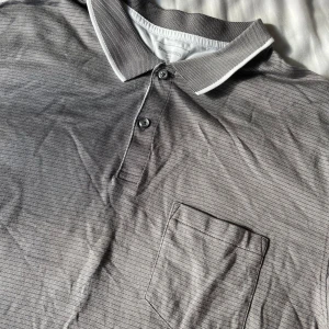 Grå pikétröja från Dressmann - Säljer en stilren grå pikétröja från Dressmann i storlek XXL. Tröjan har en klassisk krage med knappar och en praktisk bröstficka. Perfekt för både vardag och lite mer uppklädda tillfällen.