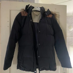 Svart pufferjacka från H&M - Säljer en svart pufferjacka från H&M:s L.O.G.G.-kollektion. Jackan har en mysig fuskpäls på huvan och bruna detaljer på axlarna. Perfekt för kalla vinterdagar med sin quiltade design och långa ärmar. Stängs med dragkedja och knappar framtill.