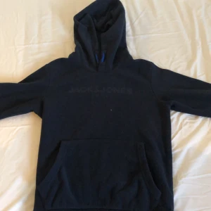 Svart hoodie från Jack & Jones - Säljer en snygg och bekväm svart fleece hoodie från Jack & Jones. Den har en klassisk design med en stor ficka fram och en huva med blå detaljer. Perfekt för en avslappnad stil!