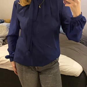 Mörkblå blus med knytband - Säljer en elegant mörkblå blus med långa ärmar och knytband vid halsen. Perfekt för både vardag och festliga tillfällen.