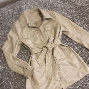Beige trenchcoat med bälte - Säljer en snygg beige trenchcoat i storlek L-XL. Jackan har långa ärmar och ett bälte i midjan för en fin passform. Den har knappar framtill och praktiska fickor på bröstet.