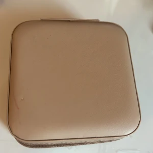 Beige smyckeskrin - Säljer ett stilrent och praktiskt beige smyckeskrin. Perfekt för att organisera dina smycken med olika fack för ringar och små krokar för örhängen. Skrinet är kompakt och lätt att ta med sig på resor. Det är i gott skick men lite smutsig 