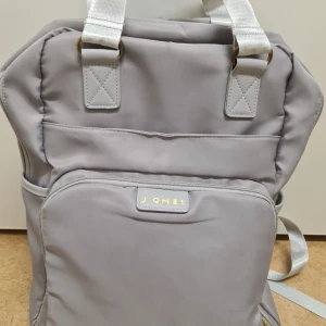 Grå ryggsäck från Jomel - Säljer en praktisk grå ryggsäck med många fickor. Den har flera fack och dragkedjor för enkel organisering. Insidan är ljus och har nätfickor för organisering. Perfekt för skolan eller jobbet!