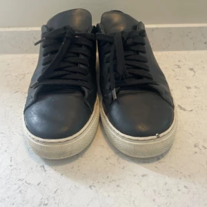 Svarta sneakers från Axel Arigato - Säljer ett par svarta sneakers från Axel Arigato. De är i skinn och har en vit sula som är lite sliten. Skorna har snörning och en klassisk design, perfekt för vardagsbruk. De är i välanvänt skick men fortfarande snygga och bekväma. Nypris 2400 kr. 