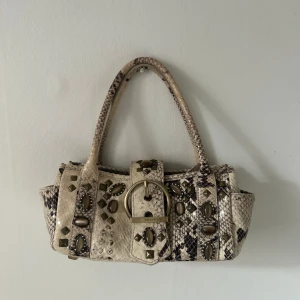 Ormmönstrad handväska från Karen Millen - Snygg ormmönstrad handväska från Karen Millen med dekorativa stenar och ett stort spänne framtill. Väskan har två handtag och en dragkedja inuti för säker förvaring. Perfekt för att ge din outfit en edgy touch. Y2k detaljer stockholmsstil