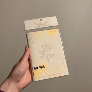 Anteckningsbok med rosdesign - En elegant anteckningsbok med ett vackert rosdesign på omslaget. Boken har en mjuk, beige ton med subtila gula inslag och texten 'ROSE' tryckt på framsidan. Perfekt för att skriva ner tankar och idéer.