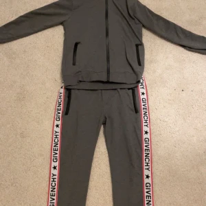 Grå Givenchy tracksuit - Snyggt grått träningsset från Givenchy med dragkedja och svarta detaljer. Byxorna har en vit och röd rand med Givenchy-logga längs sidorna. Storlek M men jag skulle säga att den är ganska  liten i storleken så S och XS passar den i också