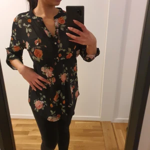 Svart blommig meshblus  - Snygg svart blus med blommigt mönster i rosa. Den har trekvartsärmar och en knytning i midjan för en smickrande passform. Perfekt för en avslappnad men stilren look.