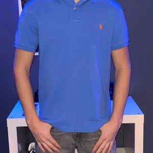 Blå pikétröja från Ralph Lauren - Säljer en snygg blå pikétröja från Ralph Lauren i slim fit. Tröjan har en klassisk krage och korta ärmar med den ikoniska broderade loggan i orange på bröstet. Perfekt för en stilren look!