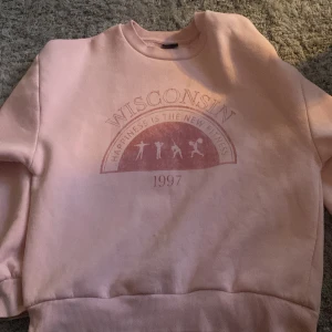 Rosa sweatshirt från Gina Tricot - Säljer en rosa sweatshirt från Gina Tricot med tryck 'Wisconsin' och 'Happiness is the new fitness 1997'. Tröjan har rund hals och ribbade muddar vid ärmslut och nederkant. Perfekt för en avslappnad stil.