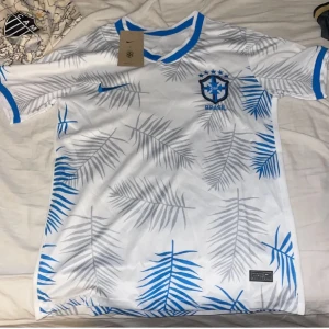 Vit och blå fotbollströja från Nike - Snygg vit fotbollströja från Nike med blå och grå palmbladsmönster. Tröjan har korta ärmar och en blå krage. Perfekt för fotbollsfans som vill sticka ut på planen!
