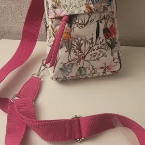 Helt ny  Blommig axelväska med rosa axelrem - Snygg axelväska med blommigt mönster och en justerbar rosa axelrem. Väskan har en dragkedja upptill och flera fack för enkel organisering. Perfekt för att ge en färgglad touch till din outfit. 22x17 cm 