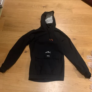 Svart anorak från Dope - Säljer en svart anorak från Dope med en stor ficka framtill och en vit logga med bergsmotiv. Anoraken har en justerbar huva med snören och långa ärmar. Perfekt för kyliga dagar i skidbacken. Skick 8/10