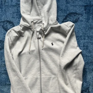 Grå hoodie från Ralph Lauren - Säljer en stilren grå hoodie från Ralph Lauren med dragkedja och klassisk logga på bröstet. Perfekt för en avslappnad look med justerbar huva och fickor framtill. Passar både till vardags och chill. Skick på tröjan 9/10 Inga söndriga grejer på tröjan 