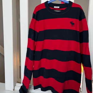 Randig tröja från Abercrombie & Fitch - Snygg långärmad tröja från Abercrombie & Fitch i rött och marinblått med breda ränder i modell Muscle. Tröjan har en broderad logotyp på bröstet. 