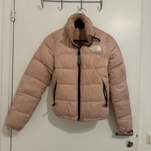 Rosa dunjacka från The North Face - Säljer en snygg rosa dunjacka från The North Face. Jackan har en dragkedja framtill och är långärmad med elastiska muddar. Perfekt för kyligare dagar.