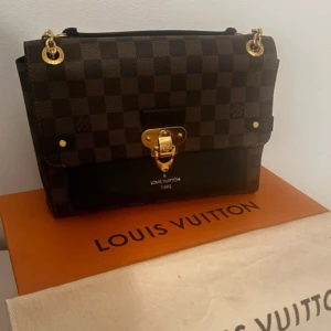 Louis Vuitton väska - Snygg brun rutig handväska från Louis Vuitton med gulddetaljer och kedjehandtag. Väskan har en elegant låsdetalj framtill och är perfekt för att ge en lyxig touch till din outfit. Köptes för 23 000kr, kvitto finns. Helt i nyskick! 