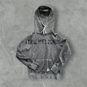 Grå hoodie från True Religion - Snygg grå hoodie från True Religion med svart text på framsidan. Den har en klassisk känguruficka och justerbar huva med snören. Perfekt för en avslappnad stil. Riktigt unik och skönt material.