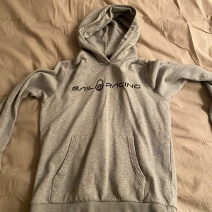 Grå hoodie från Sail Racing - Säljer en grå hoodie från Sail Racing med tryck på bröstet. Tröjan har en klassisk känguruficka och en bekväm huva. Perfekt för en avslappnad stil.