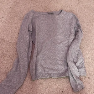 Grå stickad tröja - Säljer en mysig grå stickad tröja med långa ärmar och ribbade muddar vid ärmslut och nederkant. Perfekt för kyligare dagar.
