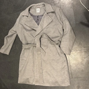 Grå kappa från Saint Tropez - Snygg grå kappa från Saint Tropez med knytbälte i midjan. Perfekt för höst och vår, med en stilren design och långa ärmar. Kappan har en klassisk krage och är tillverkad i ett mjukt material. Aldrig använd.
