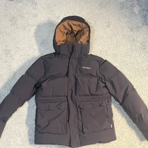 Marin blå pufferjacka från Les Deux - Snygg marin blå pufferjacka från Les Deux med brun insida. Jackan har en huva och dragkedja framtill. Den är vindtät, vattentät och andas bra, perfekt för kyliga dagar. Sprillans ny så aldrig använd, säljer pga att den är för stor. Ord pris 4500, pris kan diskuteras, 